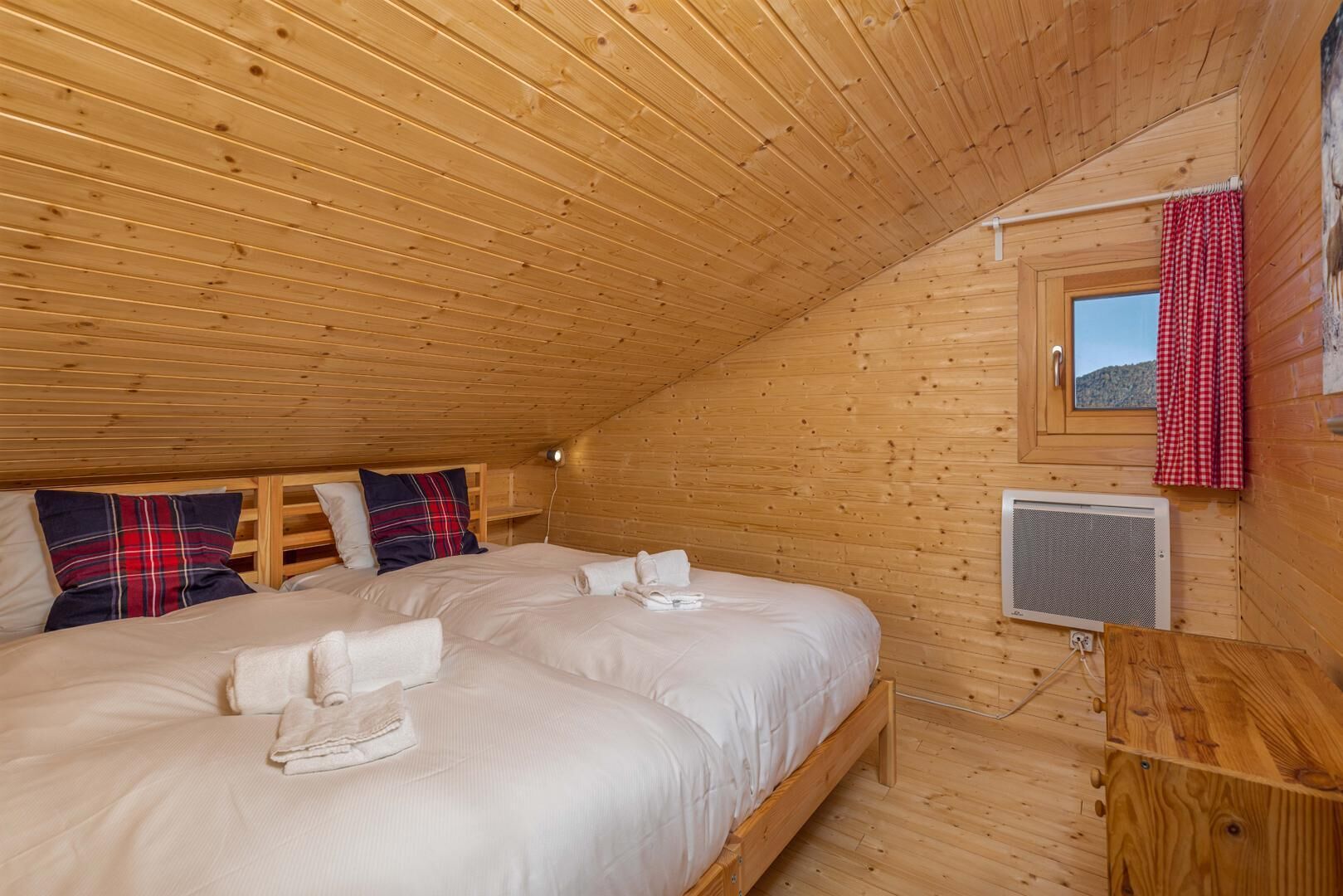 A cozy attic bedroom with wooden walls and ceiling, two beds with plaid bedding, a window with a red checkered curtain, and a wall-mounted heater.