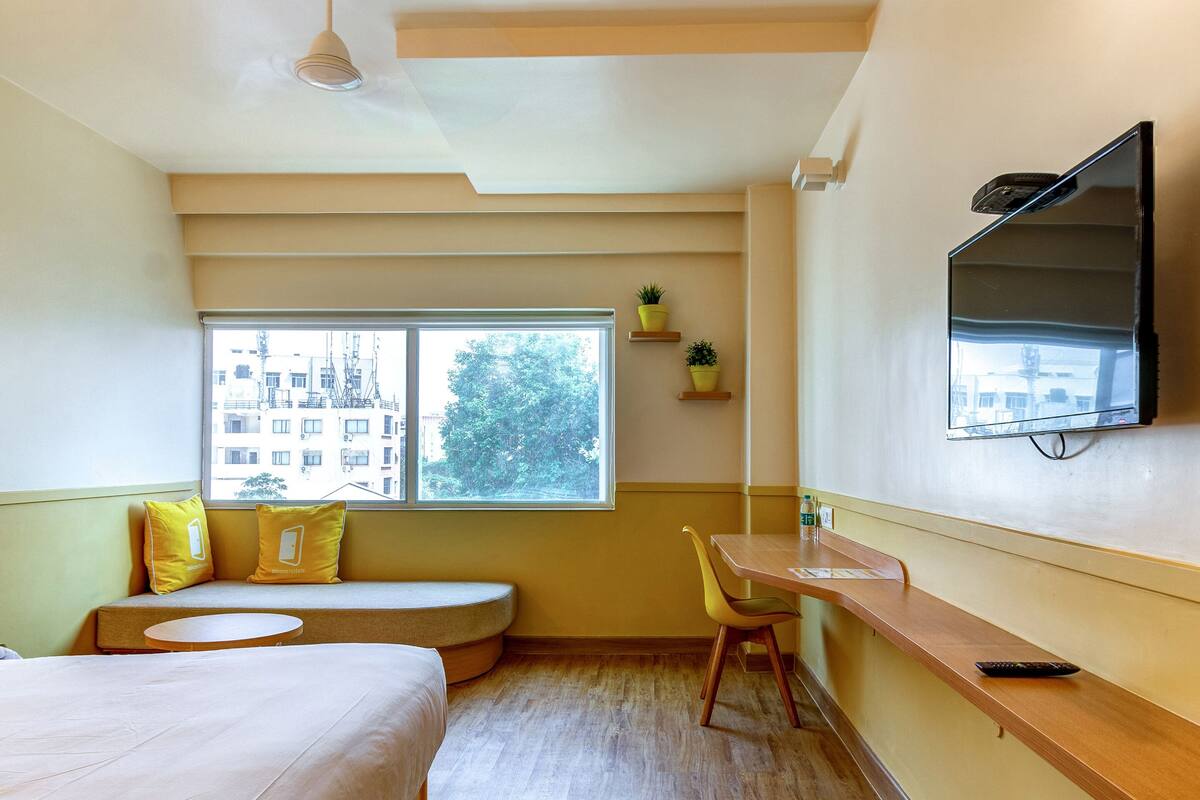 A compact hotel room with a bed, a small desk, a chair, a TV, and a window with a view of buildings and trees.