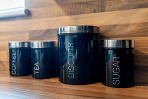Four black containers with silver lids labeled "COFFEE," "TEA," "BISCUITS," and "SUGAR" on a wooden surface.