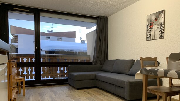 A modern living room with a grey sofa, wooden dining table, and chairs. A balcony with a glass door and a view of buildings is visible.
