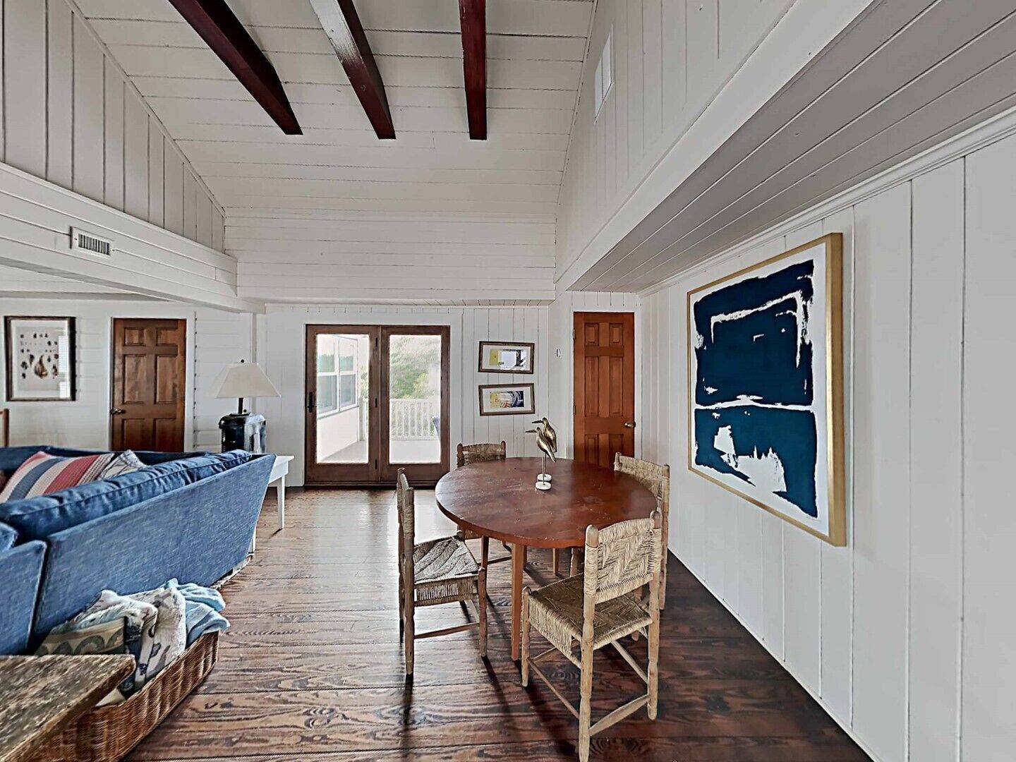 Ein Zimmer mit Holzboden, einem blauen Sofa, einem Esstisch mit Stühlen und einem großen abstrakten Gemälde an der Wand.