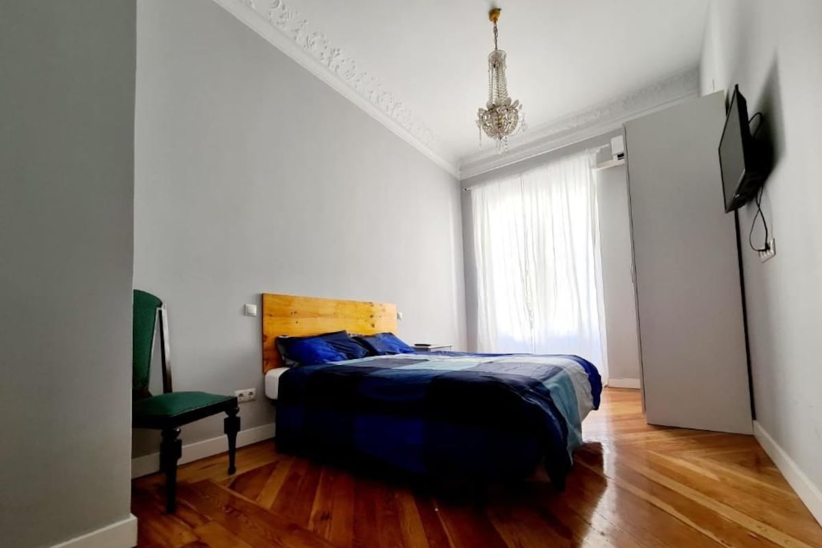 A bedroom with a wooden floor, a bed with a blue and white duvet, a green chair, a wooden headboard, a chandelier, and a TV mounted on the wall.