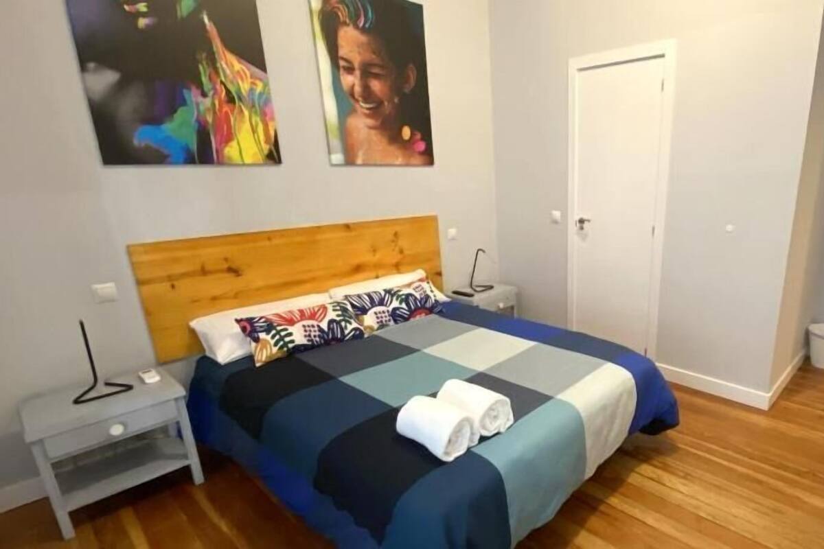 Quarto com cabeceira de madeira, colcha xadrez, mesa de cabeceira com abajur e uma porta.