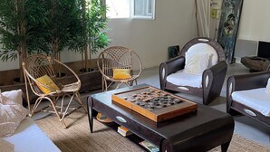 Ein Wohnzimmer mit einer Couch, zwei Sesseln, einem Couchtisch mit einem Gesellschaftsspiel, einer großen Topfpflanze und einem Bild an der Wand.
