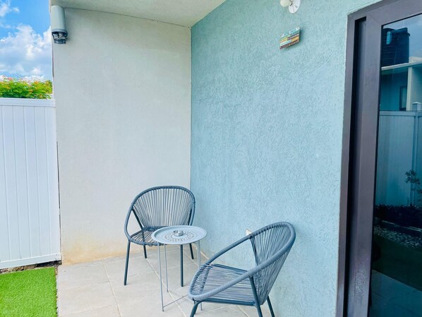 Une petite terrasse avec deux chaises en métal et une table ronde, une clôture blanche et un mur bleu à la texture particulière.