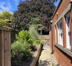 A narrow pathway leading to a brick building with a wooden fence on one side and a variety of plants and trees on the other.