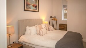 Un lit bien fait, avec une tête de lit, une lampe de chevet, une table de chevet en bois, un porte-manteau mural et une fenêtre donnant sur un immeuble.