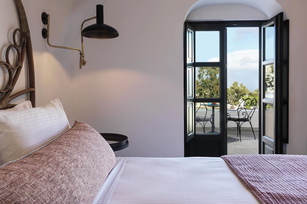 A bedroom with a bed, a bedside table, a lamp, and an open door leading to a balcony with a table and chairs.