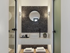 Un baño moderno con un espejo redondo, baldosas negras con motas, un lavabo blanco y dos toallas sobre una superficie de madera.