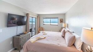 Een slaapkamer met een bed, een televisie, een raam met uitzicht op het strand en een dressoir.
