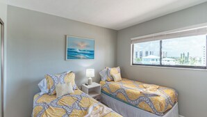 Een hotelkamer met twee bedden, een schilderij van de zee, een raam met uitzicht op de stad en een nachtkastje met een lamp.