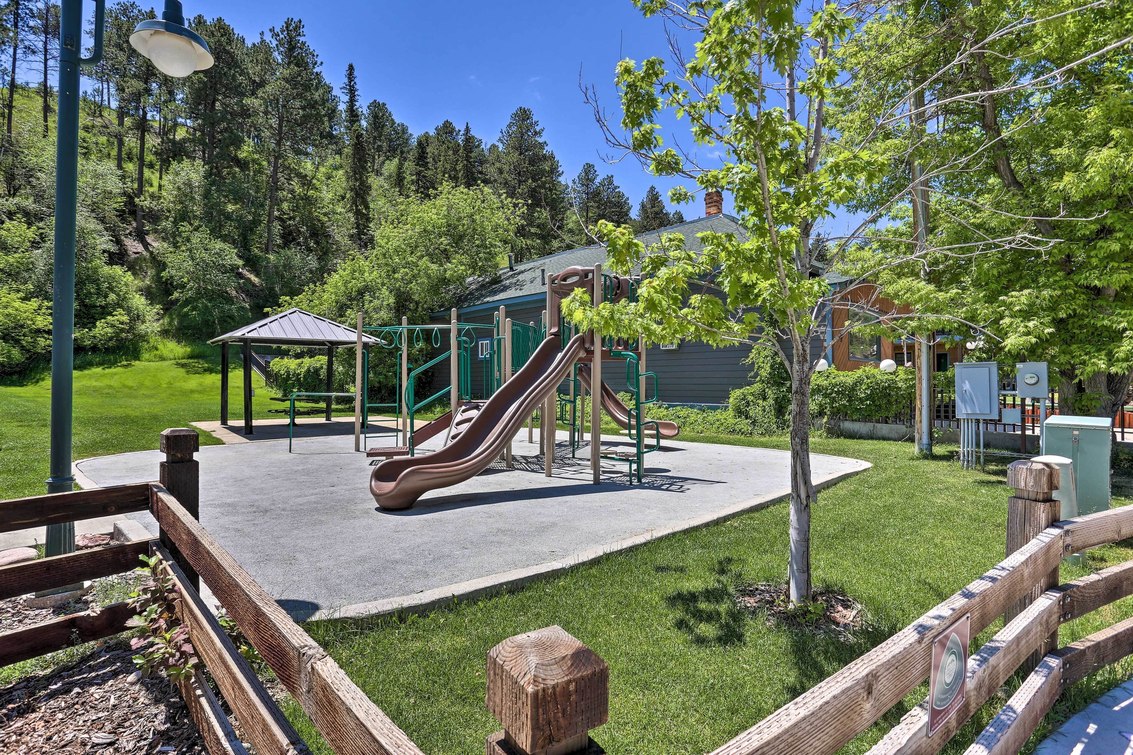 Une aire de jeux avec des toboggans et un petit abri, entourée d’herbe et d’arbres.