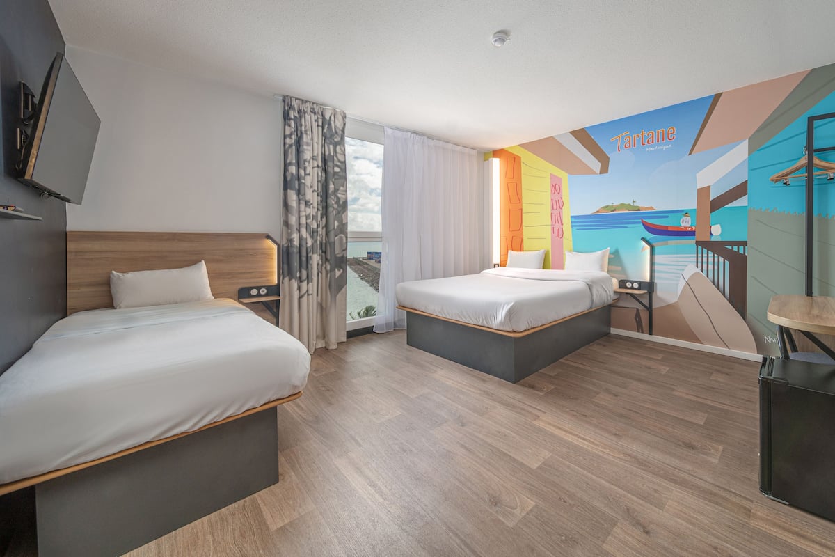 Une chambre d’hôtel avec deux lits, une grande fenêtre avec des rideaux, une fresque représentant une scène de plage et une petite table avec une chaise.