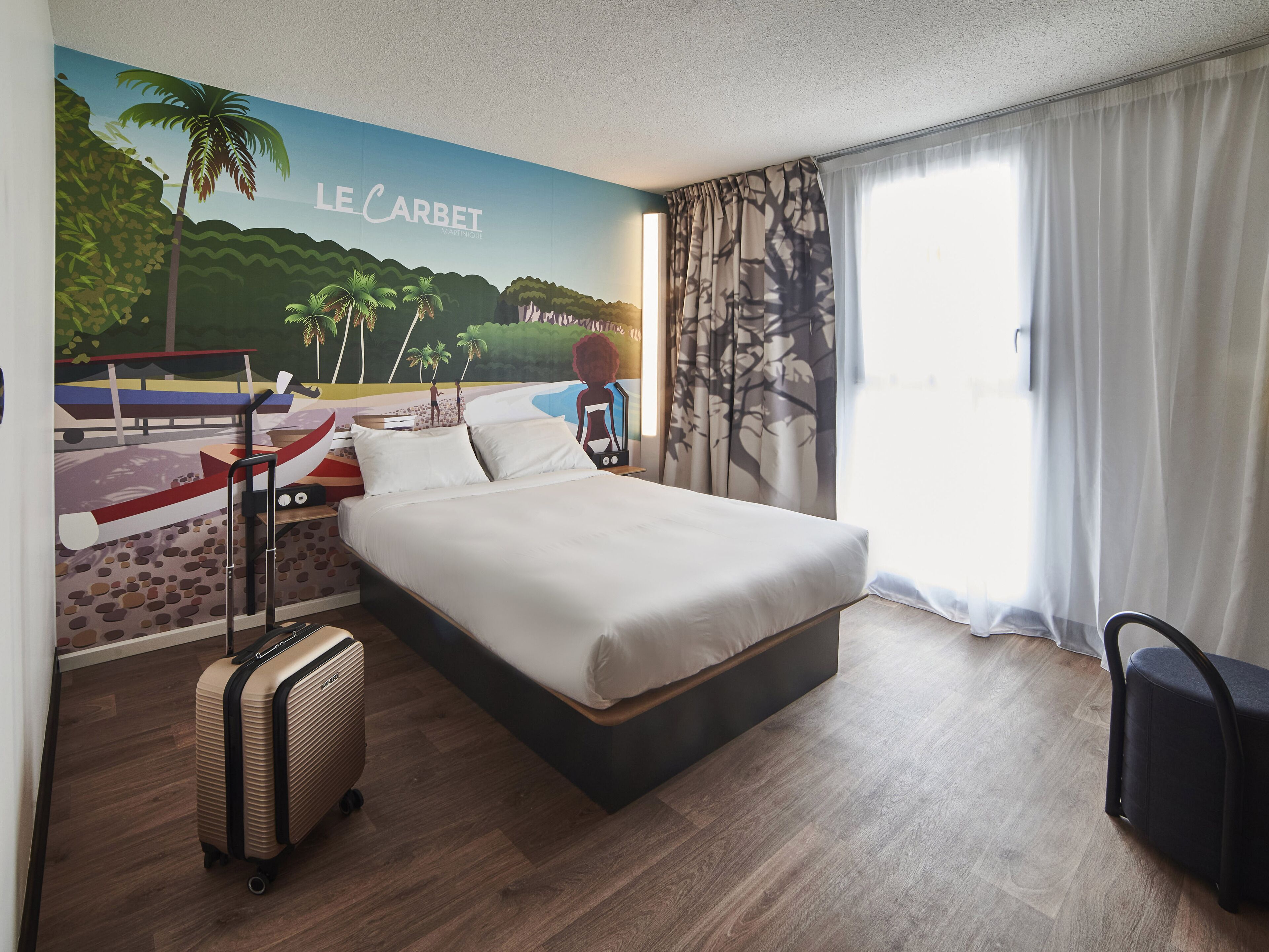 Una habitación de hotel con una cama, una maleta, una mesita de noche y un mural que representa una playa.