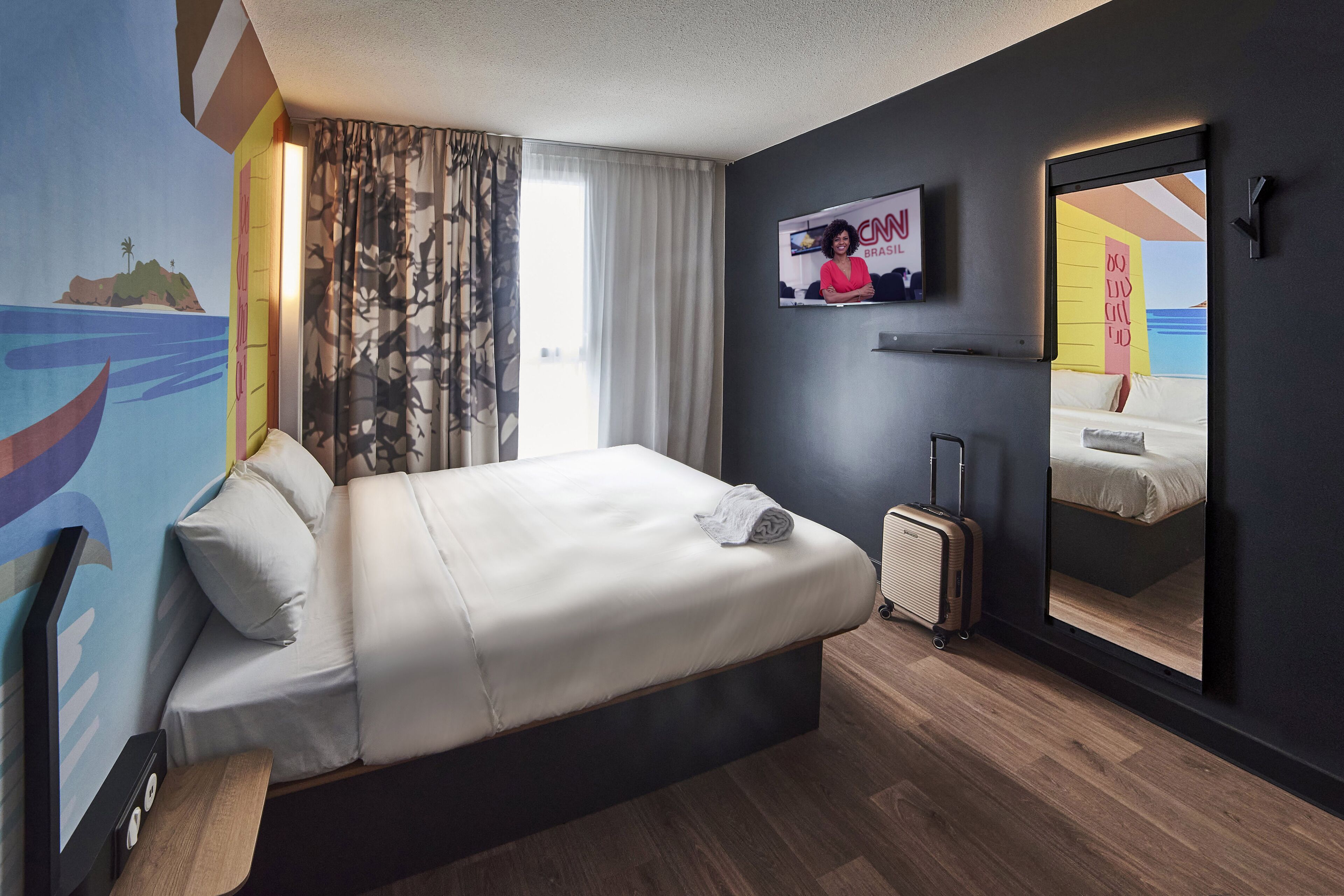 A modern hotel room with a large bed, a television, a mirror, and a window with curtains.