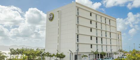 A multi-story hotel with a flat facade, a parking lot with several cars, and a clear sky with clouds.