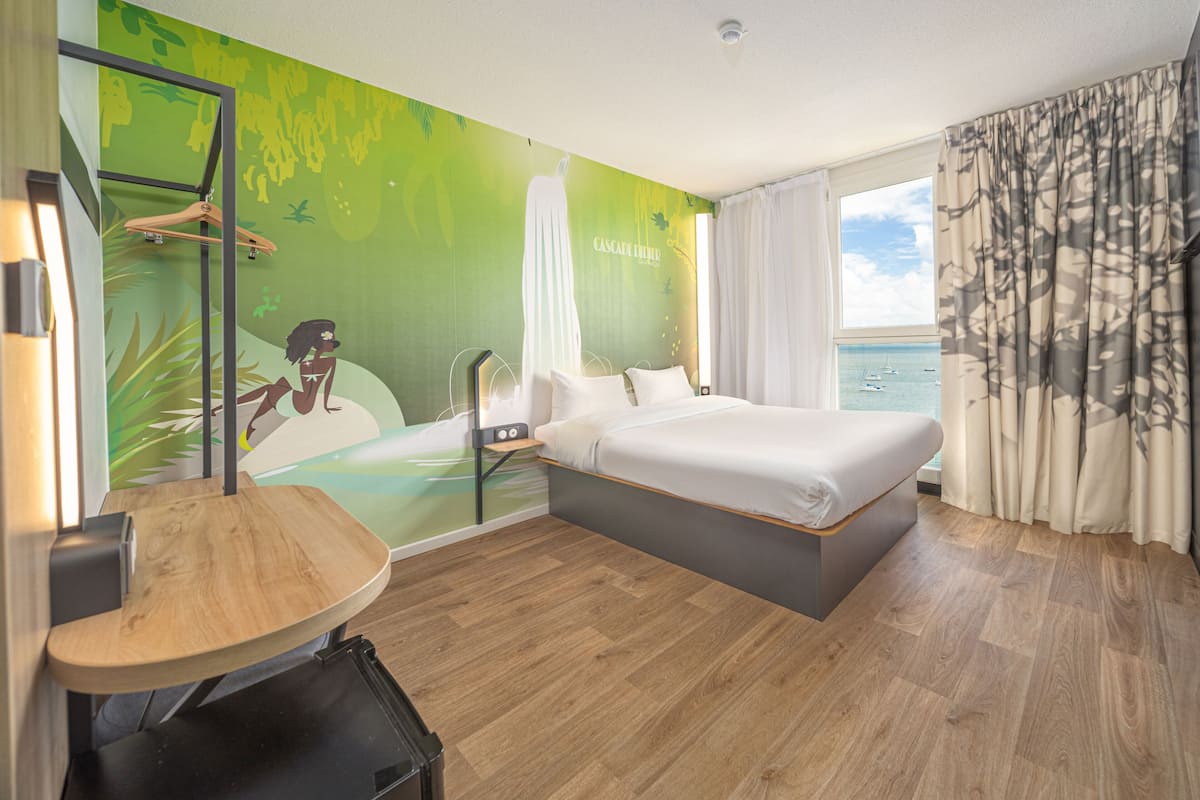 Une chambre d’hôtel comprenant un lit, un bureau, une chaise, une armoire et une grande fenêtre donnant sur la mer.