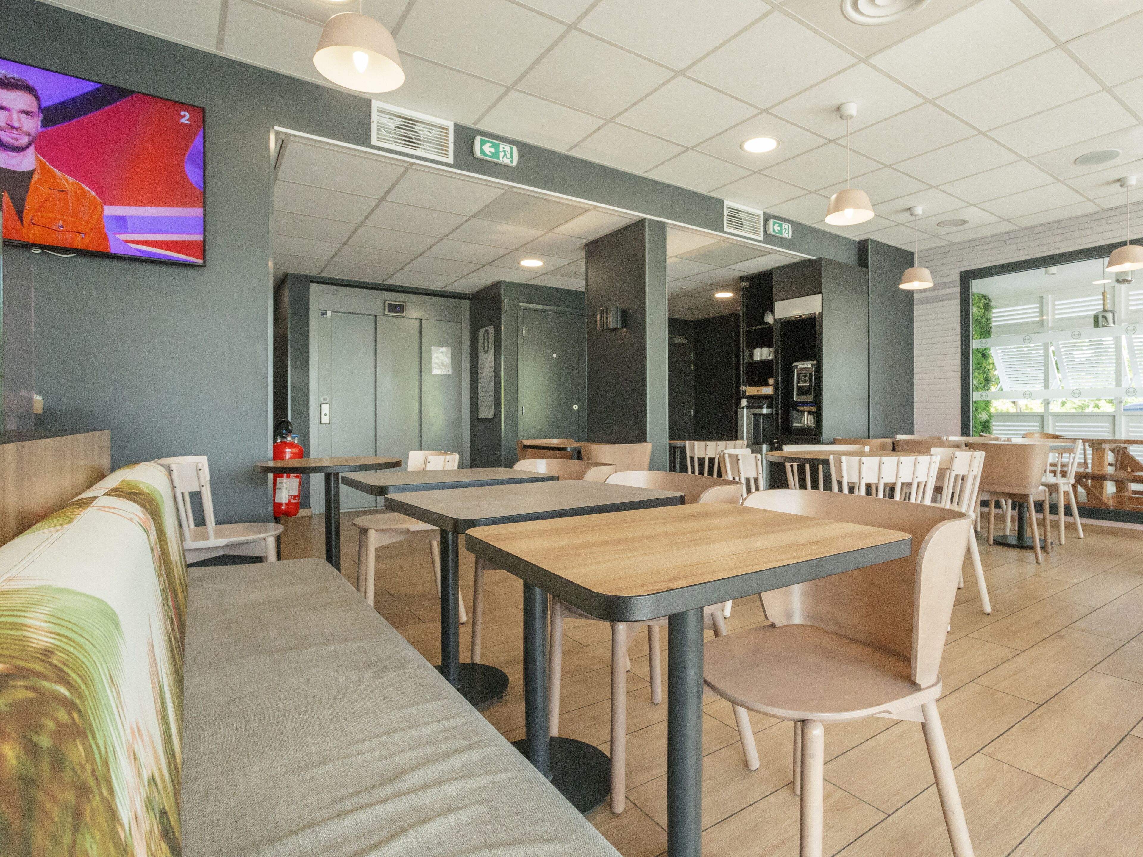 A modern restaurant with high tables and chairs, a flat-screen TV on the wall, and a fire extinguisher near the exit.