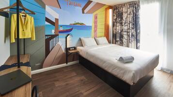 A hotel room with a bed, a desk with a laptop, a coat rack with a yellow jacket, and a mural depicting a coastal scene with the word "Tartane".