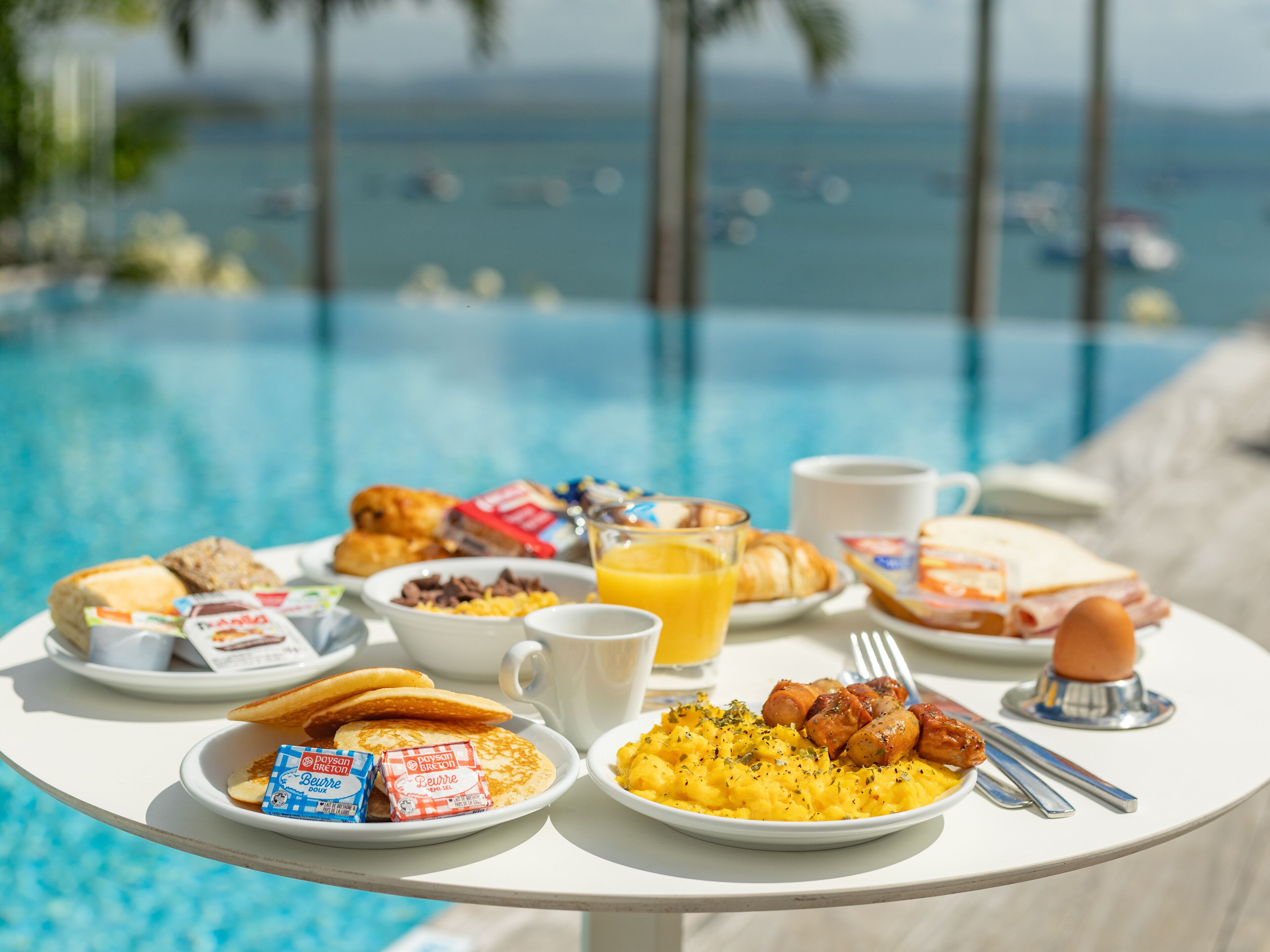 A breakfast table with croissants, pastries, and a plate of scrambled eggs by a poolside with a view of the ocean.