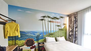 A hotel room with a bed, a desk with a chair, a suitcase, and a mural on the wall depicting a tropical scene with palm trees and a beach.