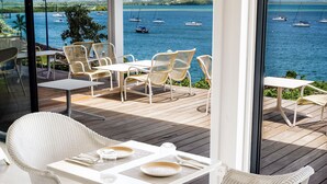 Un espace extérieur moderne, aménagé avec des chaises en osier et une table blanche, offrant une vue sur une marina avec des bateaux et une mer d’un bleu limpide.