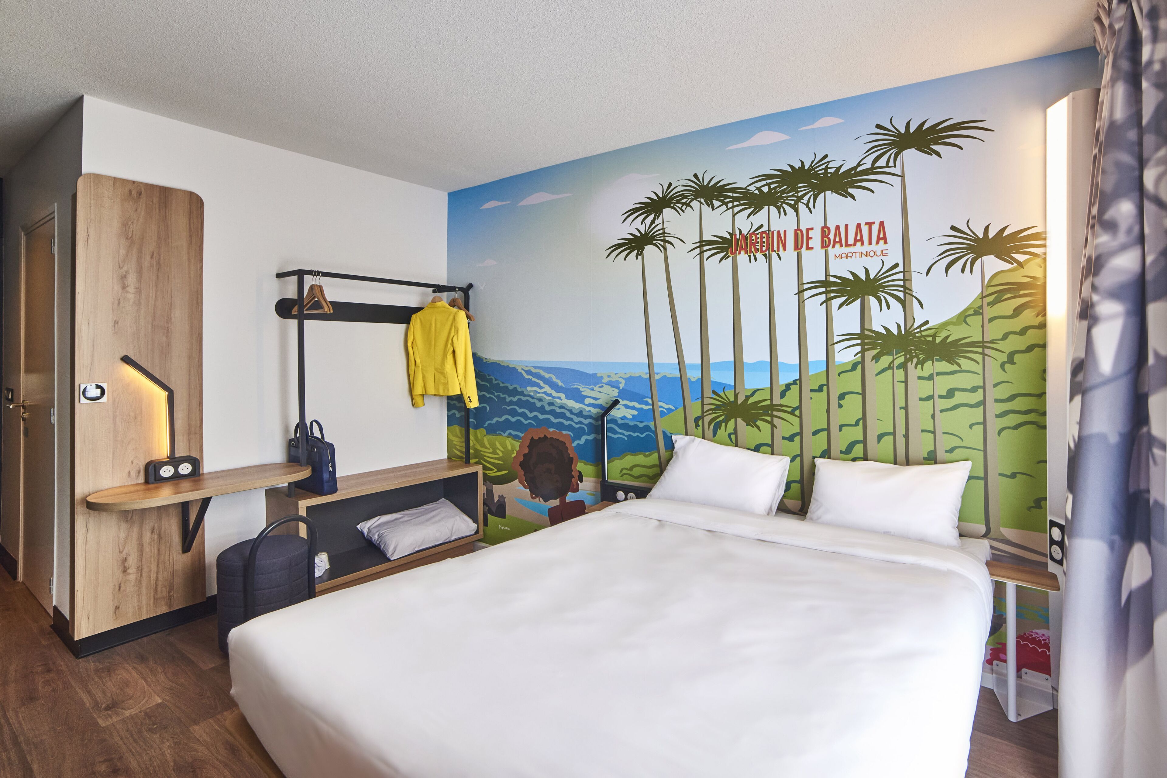 A hotel room with a bed, a wooden desk, a hanging coat rack, a wall mural, and a window with curtains.