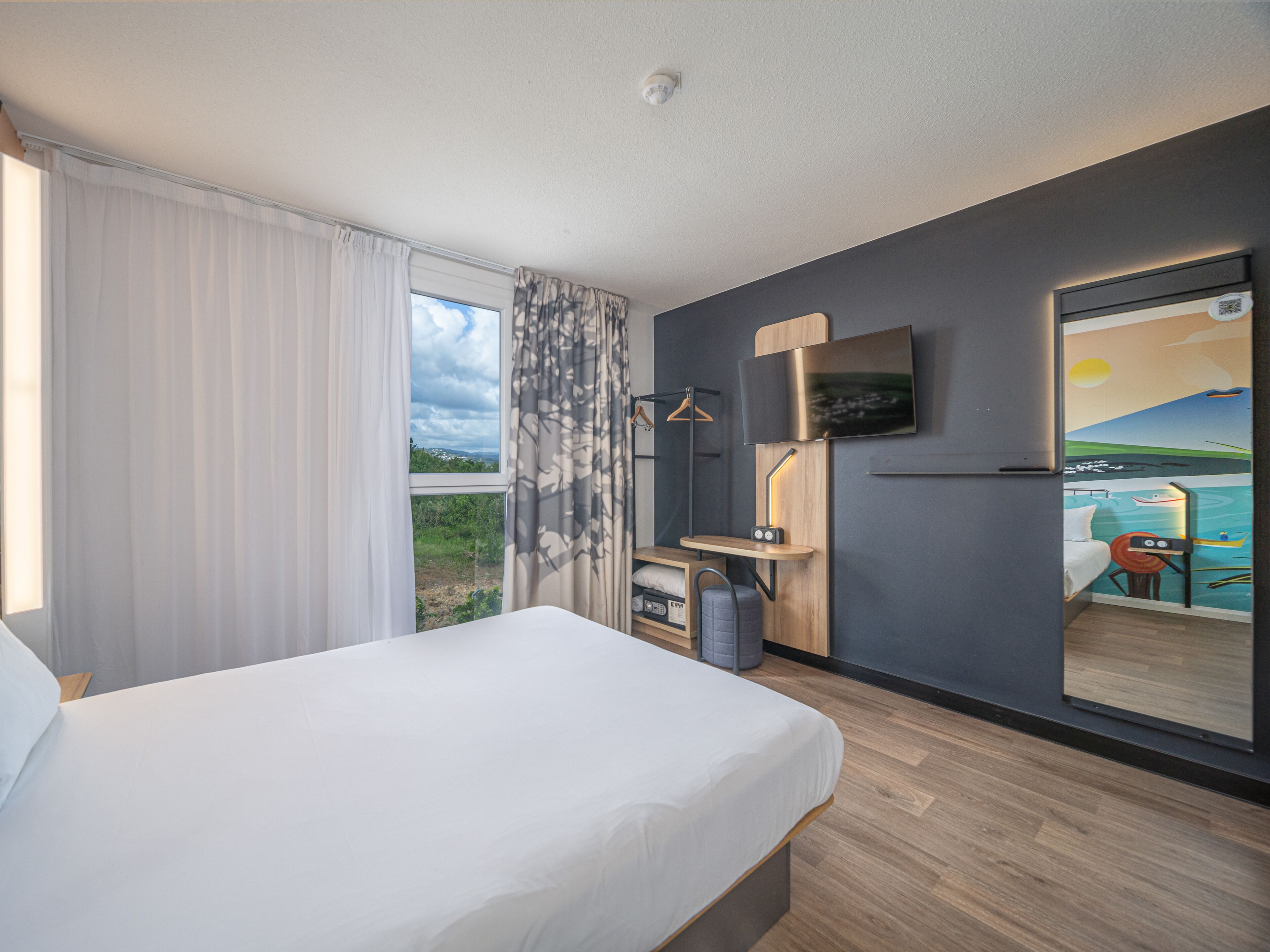 Habitación de hotel con cama, escritorio con silla, televisor montado en la pared y puerta corrediza que da a un balcón con vistas al mar.