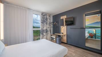 Habitación de hotel con cama, escritorio con silla, televisor montado en la pared y puerta corrediza que da a un balcón con vistas al mar.