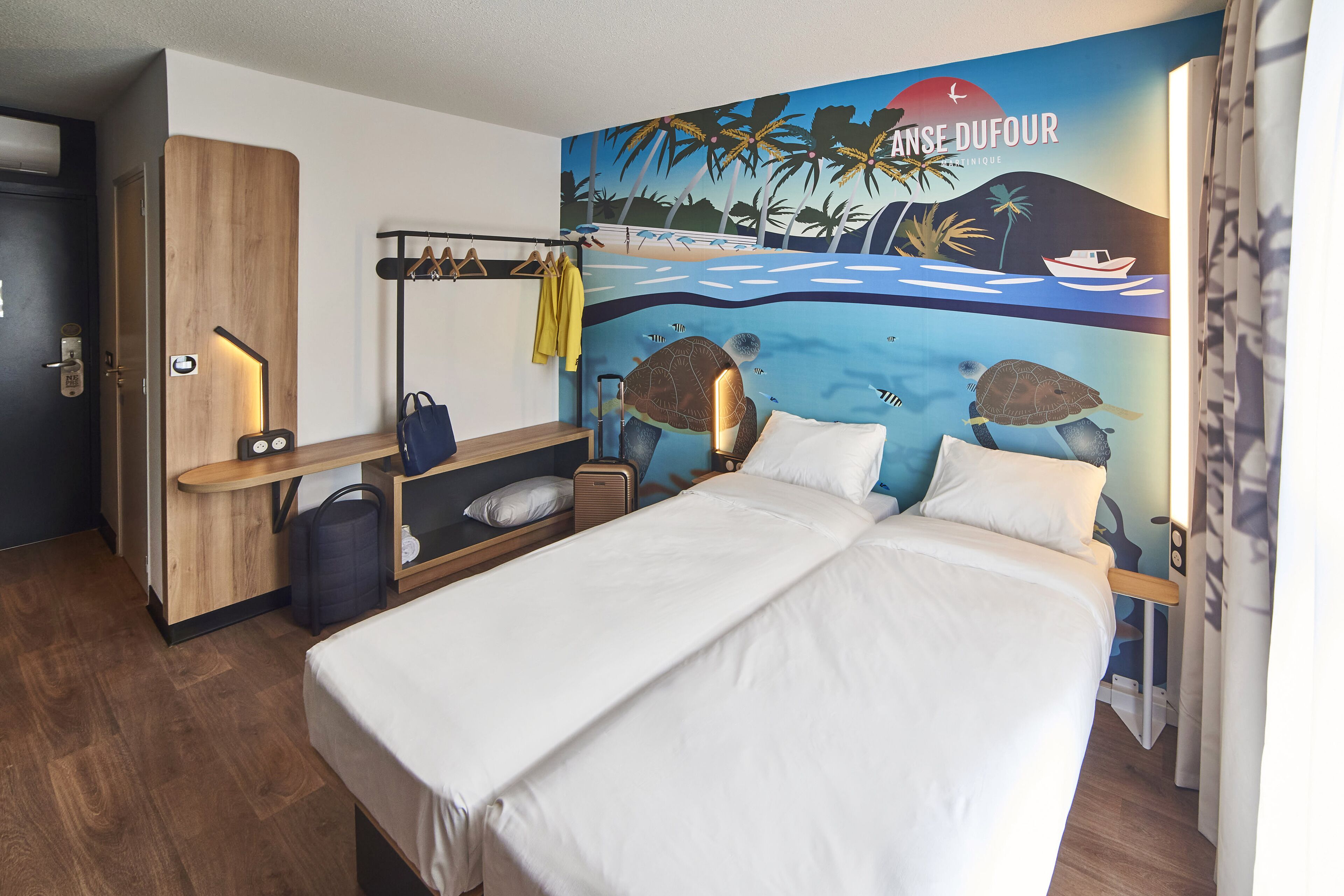 Une chambre d’hôtel avec un lit, une armoire en bois, un porte-manteau avec des vestes, une valise et une fresque murale représentant une scène de plage.