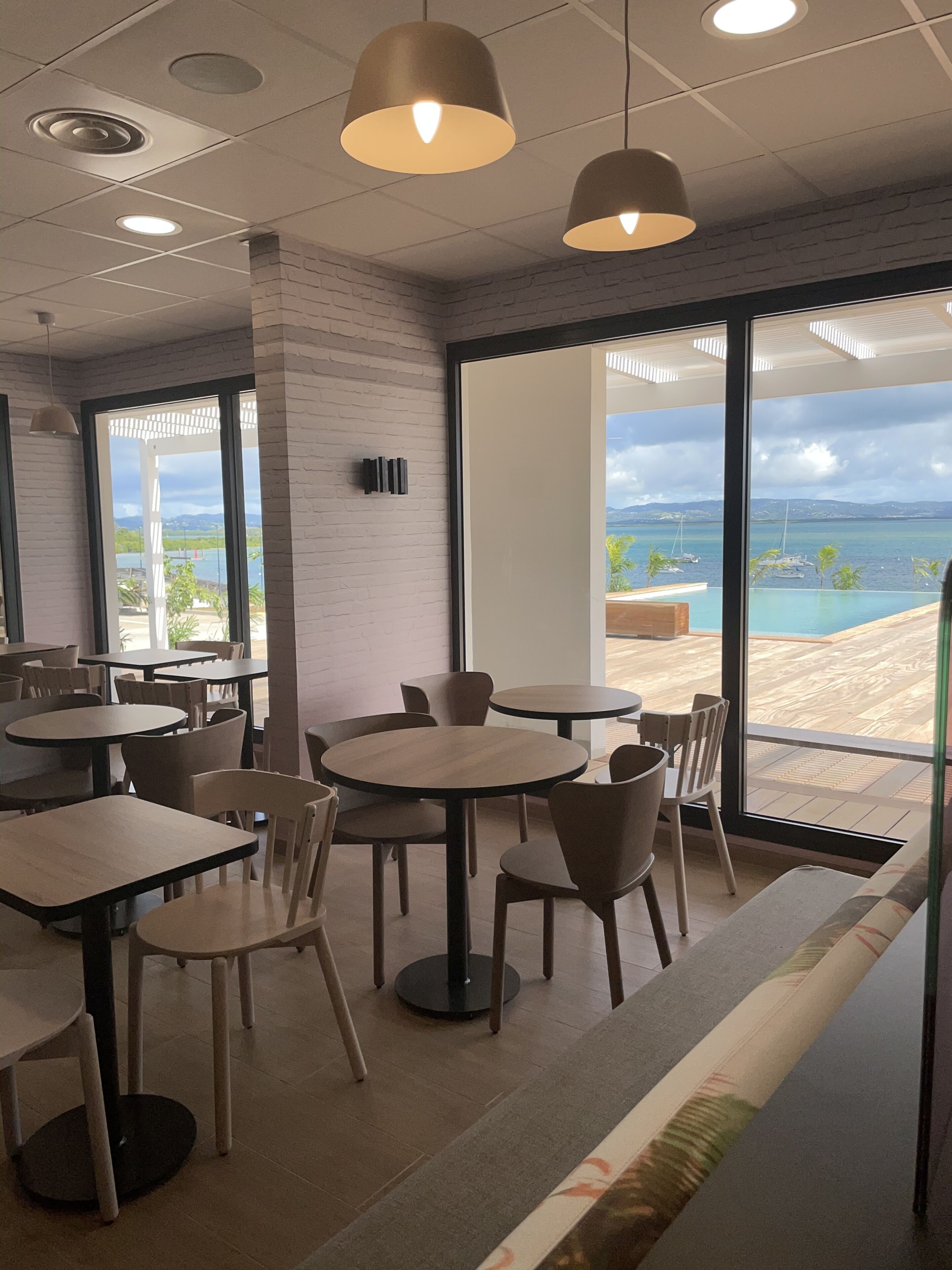 Un moderno comedor con mesas y sillas redondas, ventanas amplias con vista a una playa y al océano, e iluminación colgante.
