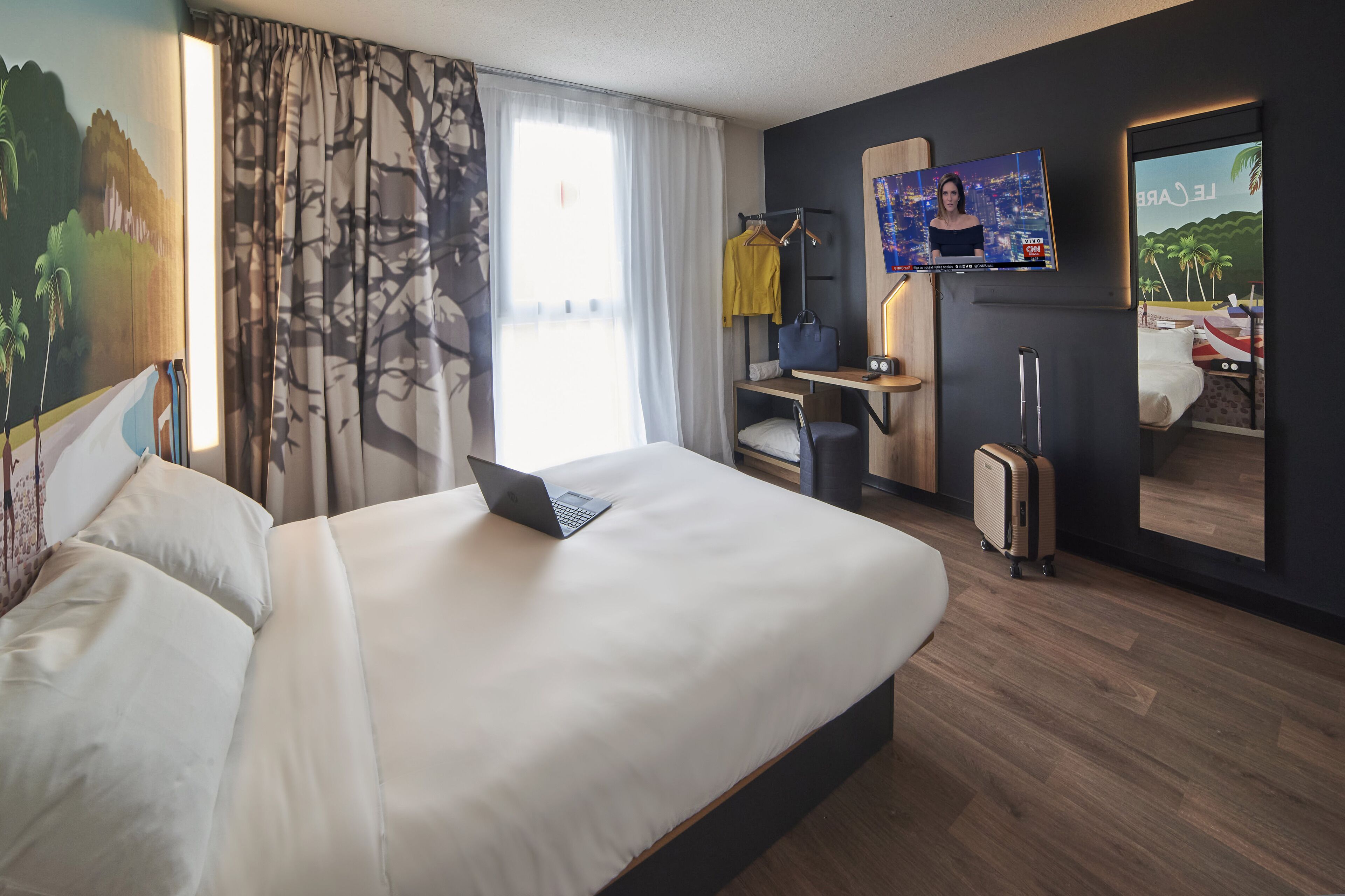 A hotel room with a bed, a desk with a laptop, a suitcase, and a TV mounted on the wall.
