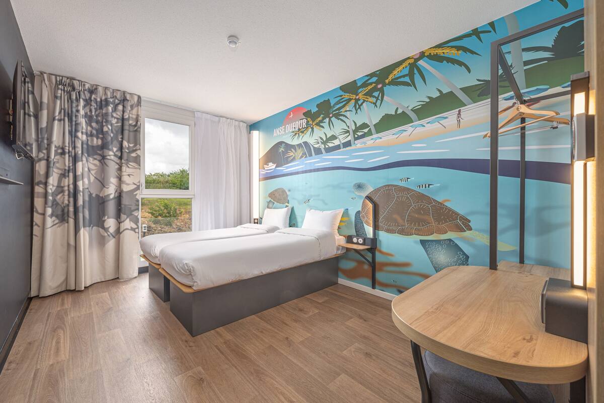 Une chambre à coucher comprenant un lit, une petite table, une chaise, un téléviseur et une fresque murale représentant une scène de plage.
