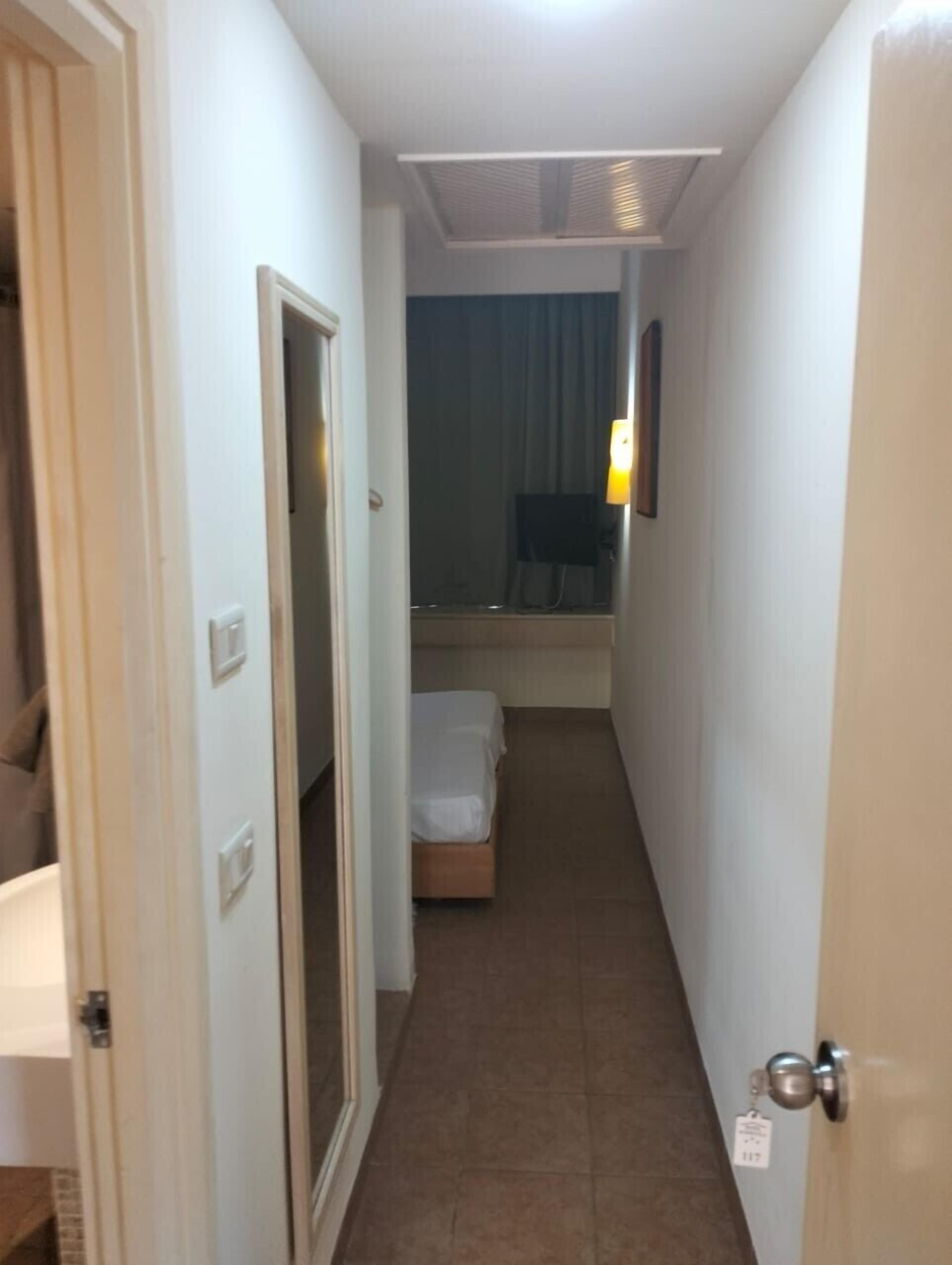 A narrow hallway with a bed at the end, a door on the left, and a light fixture above the bed.