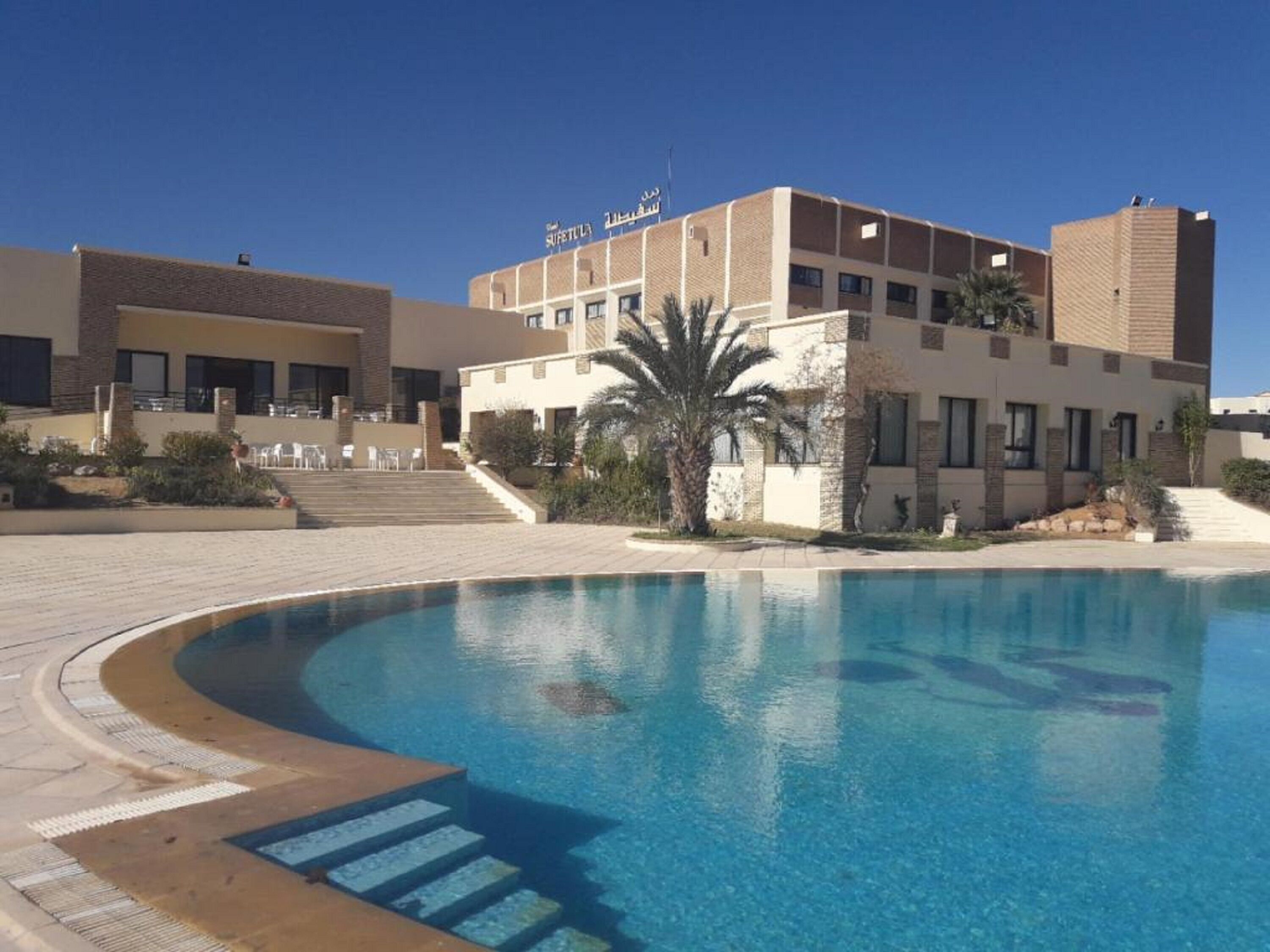 Et hotel med swimmingpool, palmetræ og trappe til indgangen.