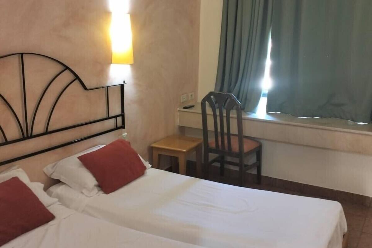 Quarto de hotel com duas camas, uma mesa de escritório, uma cadeira, janela com cortinas e luminária fixada na parede.