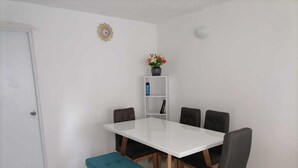 Een eethoek met een witte tafel, vier stoelen, een blauwgroene bank, een witte plank met boeken en een klok aan de muur.