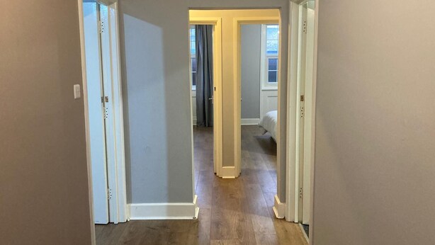 A hallway with wooden flooring and white trim, leading to a bedroom with a bed and a window. - Three Bed Two Bath Apartment in London (London)
