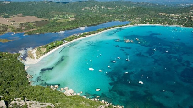 An aerial view of a coastal area with clear turquoise waters, boats, and a mountainous backdrop.