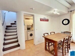 Uma cozinha compacta com máquina de lavar, uma sala de jantar com mesa e cadeiras de madeira, e uma escada que dá acesso a outro quarto.
