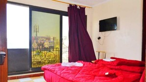 Quarto com colcha vermelha, televisão na parede e vista para uma varanda com um pôster.