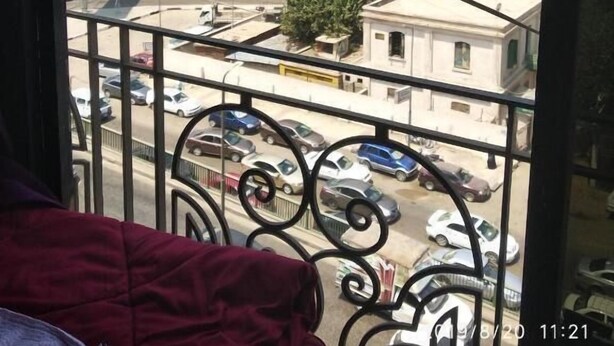 Vista de uma varanda com guarda-corrimão de metal, com vista para uma rua movimentada com carros e edifícios, incluindo um prédio alto e um outdoor.
