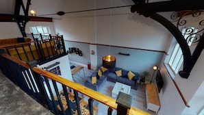 A spacious loft-style living area with a high ceiling, exposed wooden beams, and a mezzanine level with a balcony.
