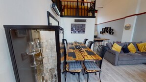 A dining area with a wooden table and chairs, a glass cabinet with wine glasses, a sofa with cushions, and a staircase with a railing.