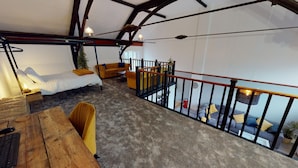 A loft-style bedroom with a bed, a desk, and a chair. There is a staircase leading to an upper level with a sofa and a plant.