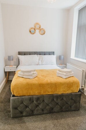 A neatly made bed with a yellow blanket and folded towels, a grey upholstered headboard, and a wall-mounted decorative piece.
