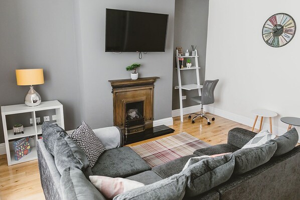 A modern living room with a grey sofa, a fireplace, a flat-screen TV, a small plant, and a white shelf with decorative items.