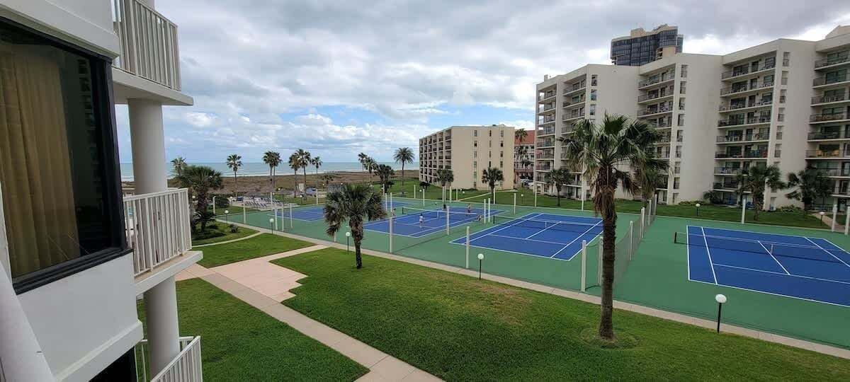 Et tennisanlæg med flere baner, omgivet af palmer og udsigt til strand og hav.