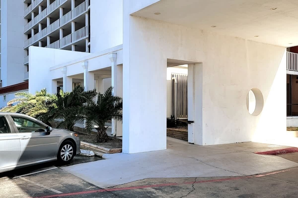 A modern building with a white facade, a car parked in front, and a small garden area with palm trees.