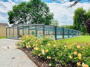 A greenhouse structure with a glass enclosure, a swimming pool, and yellow roses in the foreground. - Le Coulimachon: 2 houses on wooded land, swimming pool & water court (Méry-Bissières-en-Auge)